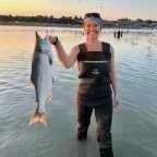 Photo of Emily Brockman standing in shallow water at sunset, smiling while holding up a large fish, with seabirds flying and people wading in the background.