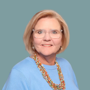Headshot of Alison Kear, smiling, with short blonde hair and clear-framed glasses, wearing a light blue top and a colorful beaded necklace against a muted teal background.