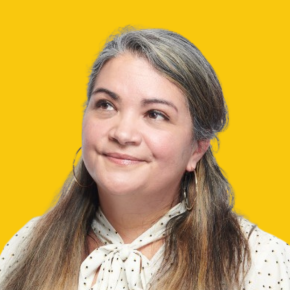 Headshot of Becca Uherbelau, looking upward with a slight smile, wearing hoop earrings and a white polka-dot blouse with a tie neck against a dandelion-yellow background.