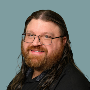 Headshot of Bill Finley, smiling, with long dark hair, glasses and a full red beard, wearing a black jacket against a muted teal background.