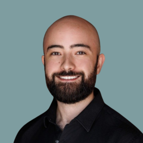 Headshot of Carlos Rojas Alvarez, smiling, with a shaved head and full dark beard, wearing a black collared shirt against a muted teal background.