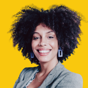 Headshot of Carolina Ramirez, smiling, with voluminous curly dark hair, a nose ring and blue geometric earrings, wearing a gray blazer against a dandelion-yellow background.