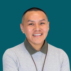Headshot of Casey Long, smiling, with a close-cropped haircut and small earrings, wearing a light gray sweater over an olive collared shirt against a dark ocean-blue background.