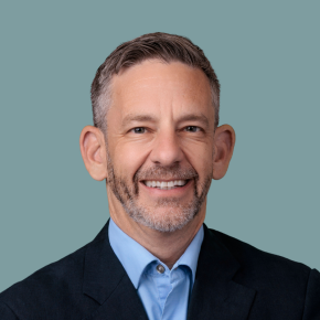 Headshot of Casey Trupin, smiling, with short salt-and-pepper hair and a trimmed beard, wearing a dark blazer over a light blue shirt against a muted teal background.