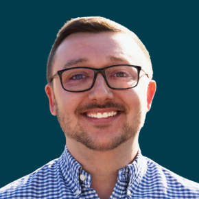 Headshot of Daniel Oxendine, smiling, with short light brown hair, glasses and a trimmed beard, wearing a blue-and-white checkered button-down shirt against a dark ocean-blue background.