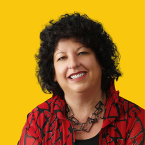 Headshot of Diane Kaplan, smiling, with short curly dark hair, wearing a red patterned jacket and black geometric necklace against a dandelion-yellow background.