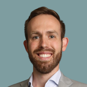 Headshot of Dylan McDowell, smiling, with short brown hair and a beard, wearing a light gray blazer over a pale blue shirt against a muted teal background.