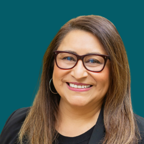 Headshot of Fabiola Greenawalt, smiling, with long light brown hair, wearing glasses, hoop earrings and a black blazer over a black top against a dark ocean-blue background.