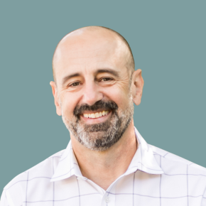 Headshot of Glen Galaich, smiling, with a shaved head and salt-and-pepper beard, wearing a white collared shirt against a muted teal background.
