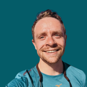 Headshot of Ian Finch, smiling outdoors, with short light brown hair and a beard, wearing a teal shirt with sunglasses hanging around his neck against a dark ocean-blue background.