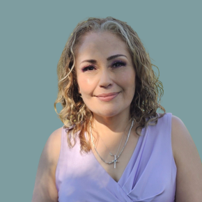 Headshot of Iris Alexandra Lopez, smiling softly, with shoulder-length curly blonde hair, wearing a lavender sleeveless top and silver necklaces against a muted teal background.