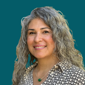 Headshot of Jackie Murphy, smiling, with long curly gray hair, wearing a black-and-white patterned shirt and a green pendant necklace against a dark ocean-blue background.