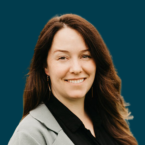 Headshot of Jamie Carroll, smiling with long brown hair and wearing a light gray blazer over a black top, against a dark teal background.