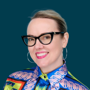 Headshot of Jean Westrick, smiling, with black cat-eye glasses, silver hoop earrings and bright pink lipstick, wearing a colorful patterned blouse against a dark ocean-blue background.