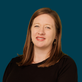 Headshot of Jeri Rathbun, looking at the camera with a slight smile, with straight red hair, wearing a black top and hoop earrings against a dark ocean-blue background.