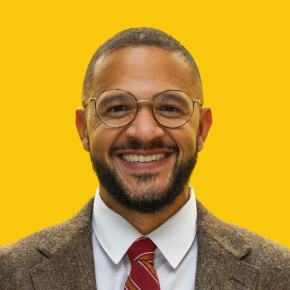 Headshot of Jesse Beason, smiling, wearing round glasses, a brown blazer, white shirt and red striped tie against a dandelion-yellow background.