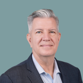 Headshot of John Mohr, looking at the camera with a slight smile, with short silver hair, wearing a dark blazer over a light blue shirt against a muted teal background.