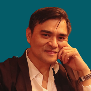 Headshot of Jose Antonio Vargas, looking at the camera with smile, with dark hair and one hand resting near his temple, wearing a black shirt against a dark ocean-blue background.