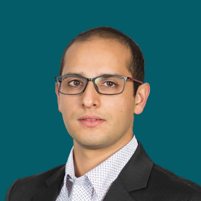 Headshot of Juan Clavijo, looking at the camera with a neutral expression, wearing glasses, a dark suit jacket, and a white patterned shirt against a dark ocean-blue background.