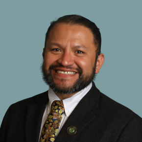 Headshot of Juan Martinez Pineda, smiling, with short dark hair and a beard, wearing a black suit jacket, white shirt, patterned tie and lapel pin against a muted teal background.