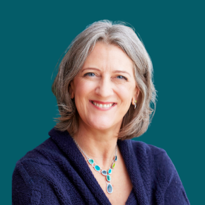 Headshot of Judith Wiencken, smiling, with shoulder-length gray hair, wearing a navy sweater and a turquoise necklace against a dark ocean-blue background.