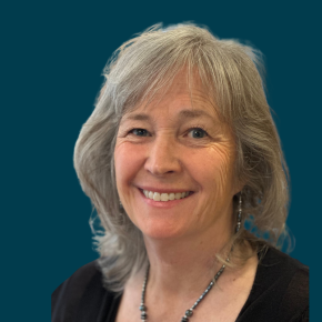 Headshot of Julie Greene Graham, smiling, with shoulder-length gray hair, wearing dangling earrings, a black top and a beaded necklace against a dark ocean-blue background.