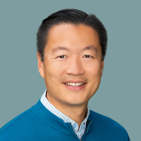 Headshot of Justin Yuen, smiling, with short black hair, wearing a teal zip sweater over a blue-and-white striped shirt against a muted teal background.
