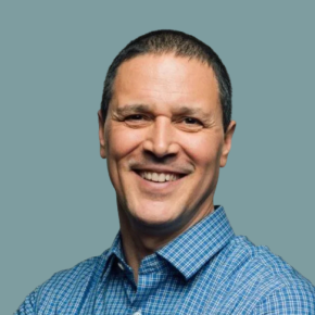 Headshot of Kameron Perez-Verdia, smiling with short dark hair and wearing a blue checkered shirt, against a muted blue-gray background.