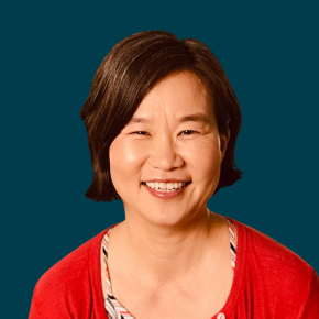 Headshot of Katie Hong, smiling, with short dark hair, wearing a red cardigan over a patterned top against a dark ocean-blue background.