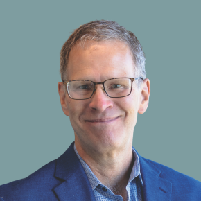 Headshot of Kevin Walker, smiling, with short gray hair and glasses, wearing a blue blazer over a patterned blue shirt against a muted teal background.