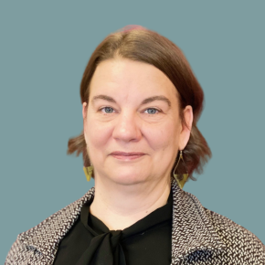 Headshot of Laura Forbes, looking at the camera with a slight smile, wearing geometric earrings, a black blouse with a tie neck, and a patterned blazer against a muted teal background.