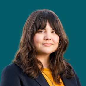 Headshot of Lauren Waudé, looking at the camera with a calm expression, with shoulder-length wavy brown hair and bangs, wearing a navy blazer over a mustard top against a dark ocean-blue background.