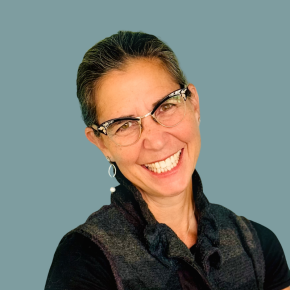 Headshot of Laurie Wolf, smiling, with dark hair pulled back, wearing patterned glasses, dangling earrings, and a dark textured vest against a muted teal background.