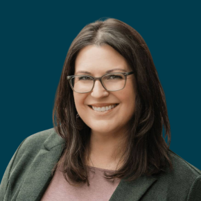 Headshot of Lee Ann Grogan, smiling, with shoulder-length dark brown hair, glasses, and a small nose ring, wearing a green blazer over a mauve top against a dark ocean-blue background.
