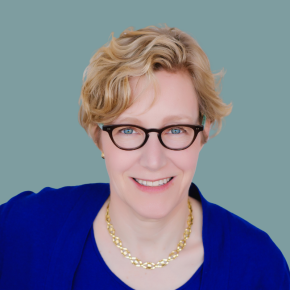Headshot of Libby Hlavka, smiling, with short curly blonde hair and black glasses, wearing a royal blue top and gold chain necklace against a muted teal background.