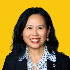 Headshot of Lin Zhou, smiling at the camera, with shoulder-length black hair, wearing a black blazer, patterned white-and-blue blouse, pearl necklace, and pearl earrings against a dandelion-yellow background.