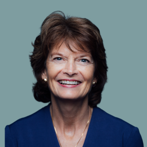 Headshot of Lisa Murkowski, smiling with short brown hair and wearing gold earrings, a gold necklace and a blue blazer, against a muted blue-gray background.