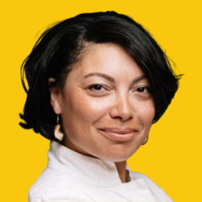 Headshot of Loki Tobin, looking at the camera with a slight smile, with short black hair swept to one side, wearing a white high-collared top and dangling earrings against a dandelion-yellow background.
