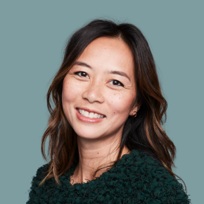 Headshot of Luan Nio, smiling, with shoulder-length wavy dark brown hair and subtle highlights, wearing a textured dark green top against a muted teal background.