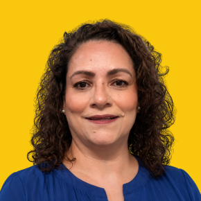Headshot of Magdalena Ochoa Gomez, looking at the camera with a soft smile, with shoulder-length curly dark brown hair, wearing pearl earrings and a royal blue blouse against a dandelion-yellow background.