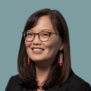 Headshot of Margie Andreason, smiling, with shoulder-length dark hair and glasses, wearing a black blazer and red-and-gold earrings against a muted teal background.