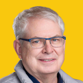 Headshot of Marty Kooistra, smiling slightly, with short white hair and blue glasses, wearing a light gray jacket against a dandelion-yellow background.