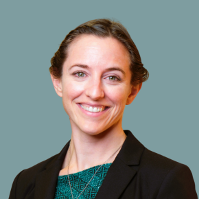 Headshot of Meadow Didier, smiling, with hair pulled back, wearing a black blazer over a teal patterned top against a muted teal background.