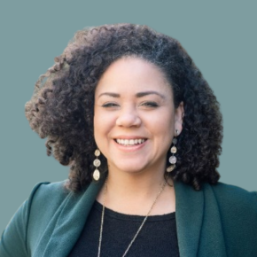 Headshot of Melissa Sines, smiling, with voluminous curly dark hair, wearing drop earrings, a necklace, and a dark green blazer over a black top against a muted teal background.