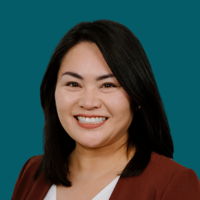 Headshot of Meredith Higashi, smiling, with shoulder-length black hair, wearing a rust-colored blazer over a white top against a dark ocean-blue background.