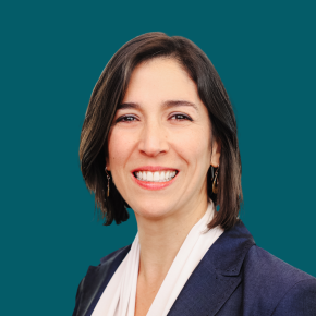 Headshot of Michelle Boss Barba, smiling, with shoulder-length dark brown hair, wearing a navy blazer over a white blouse and dangling earrings against a dark ocean-blue background.