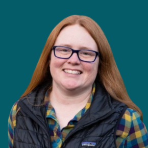 Headshot of Michelle DeWitt, smiling, with long red hair and glasses, wearing a black vest over a blue-and-yellow plaid shirt against a dark ocean-blue background.