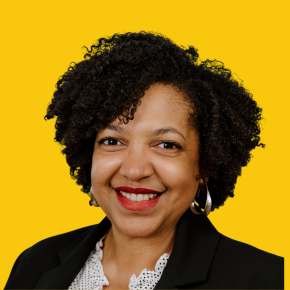 Headshot of Nancy Sanabria, smiling with short curly dark hair and wearing silver hoop earrings and a black blazer, against a bright yellow background.
