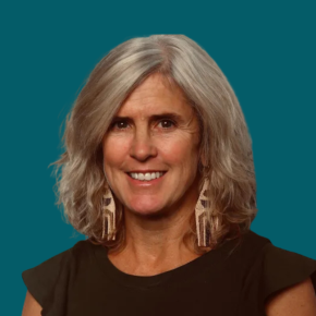 Headshot of Polly Carr, smiling with shoulder-length gray hair and wearing long beaded earrings and a black top, against a teal background.