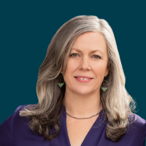 Headshot of Robin Hill-Dunbar, looking at the camera with a slight smile, with long silver-gray hair, wearing a purple blouse and green geometric earrings against a dark ocean-blue background.